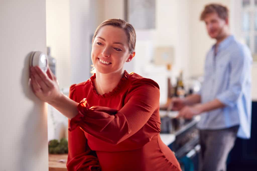 A woman turning down the thermostat while her husband looks on from the other room