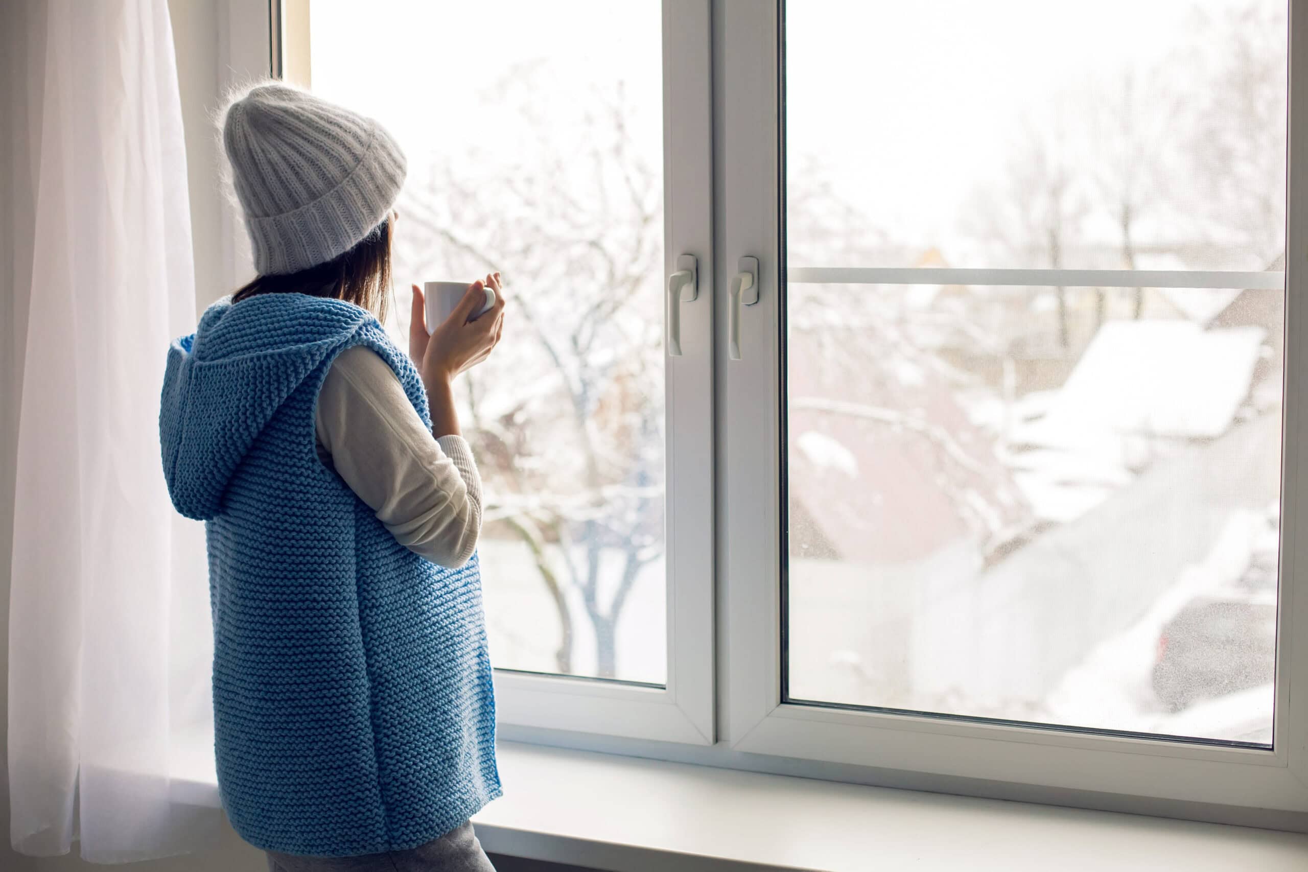 Cozy winter scene with a person in a blue sweater holding a mug, gazing out a window at a snow-covered landscape